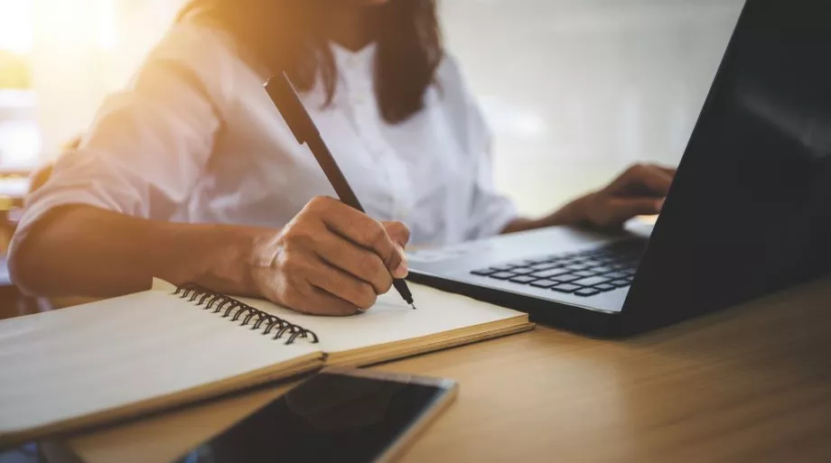 Une femme devant son ordinateur qui prend des notes sur un cahier