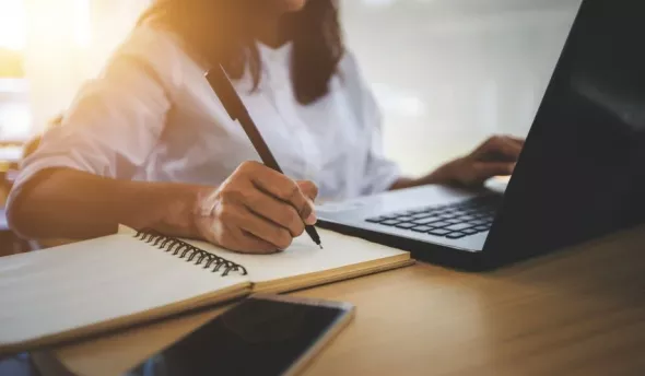 Une femme devant son ordinateur qui prend des notes sur un cahier