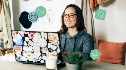 Jeune femme souriante tapant sur un ordinateur portable décoré de stickers, dans un coin de travail chaleureux.